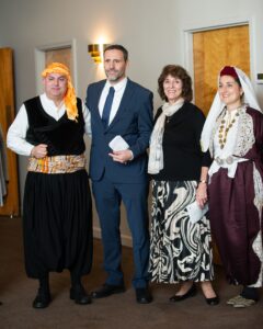 Two men and two women posing for a photograph. The man on the far left and the woman on the far right are wearing traditional Greek costumes. The man in the middle is wearing a suit and the woman in the middle is wearing formal attire.